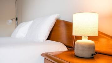 Un lit avec une literie blanche et une tête de lit en bois, une table de chevet avec une lampe à texture intéressante et un luminaire fixé au mur.