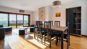 Uma área de jantar moderna com mesa e cadeiras de madeira, parede de destaque em pedra e vista através de grandes janelas.