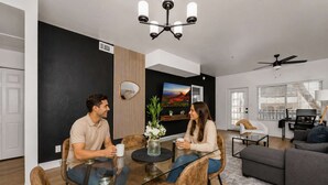 A modern living room with a dining table, two people seated, a sofa, a TV, and a ceiling fan.