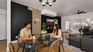 A modern living room with a dining table, two people seated, a sofa, a TV, and a ceiling fan.