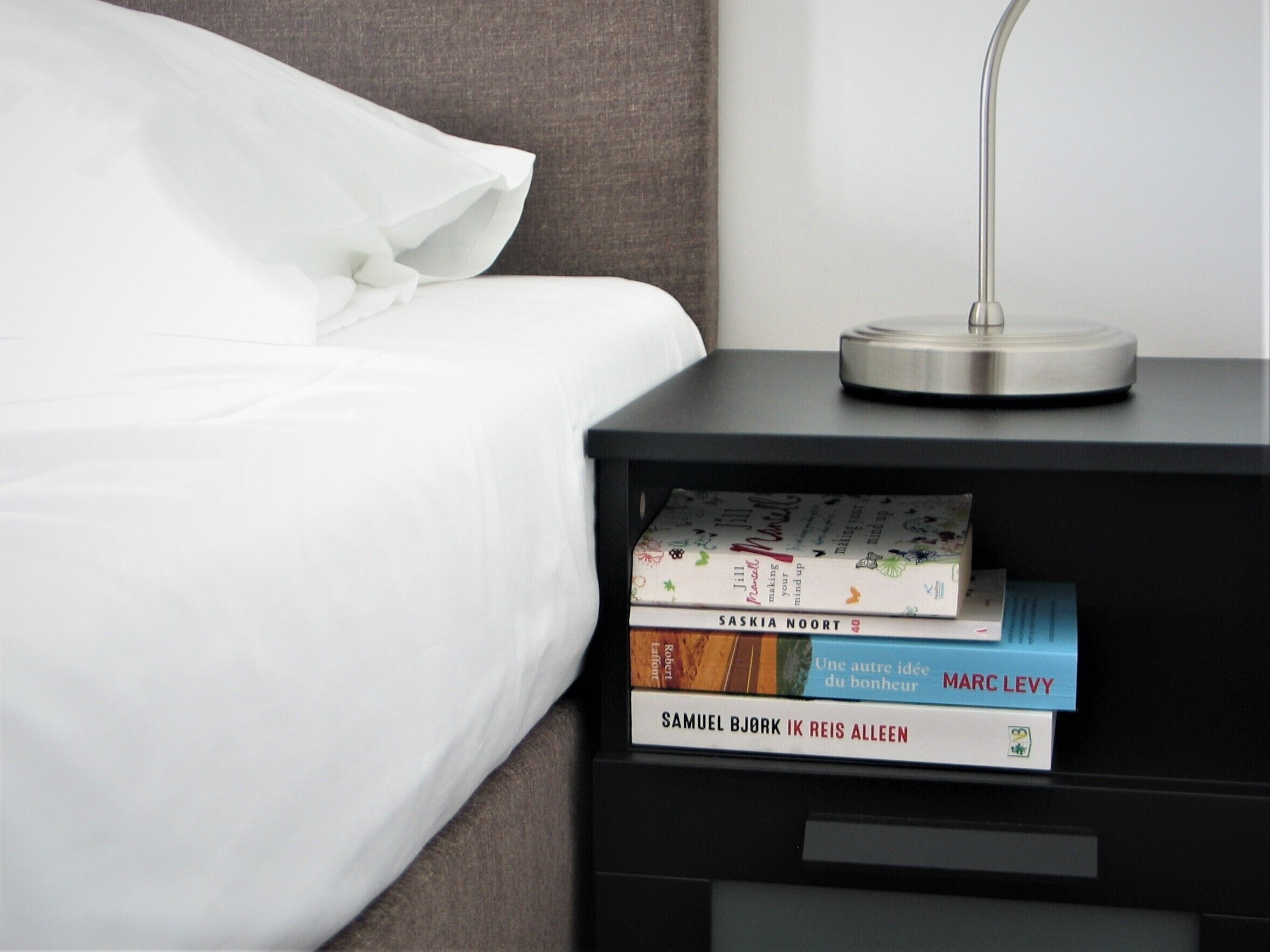 A neatly made bed with white sheets and pillows, a bedside table with a lamp, and a stack of books on the table.