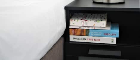 A neatly made bed with white sheets and pillows, a bedside table with a lamp, and a stack of books on the table.