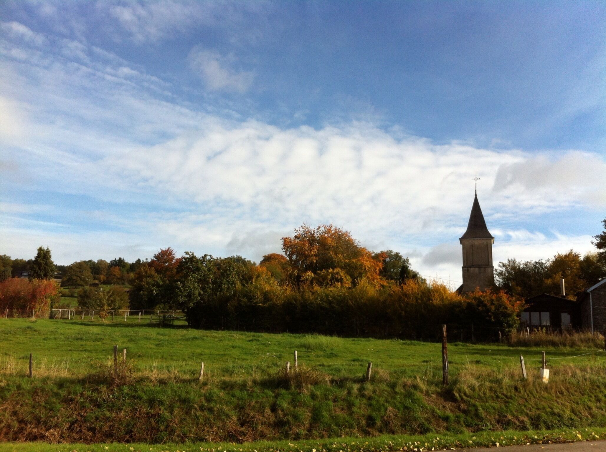 A rural landscape with a church tower, green fields, and autumn trees.