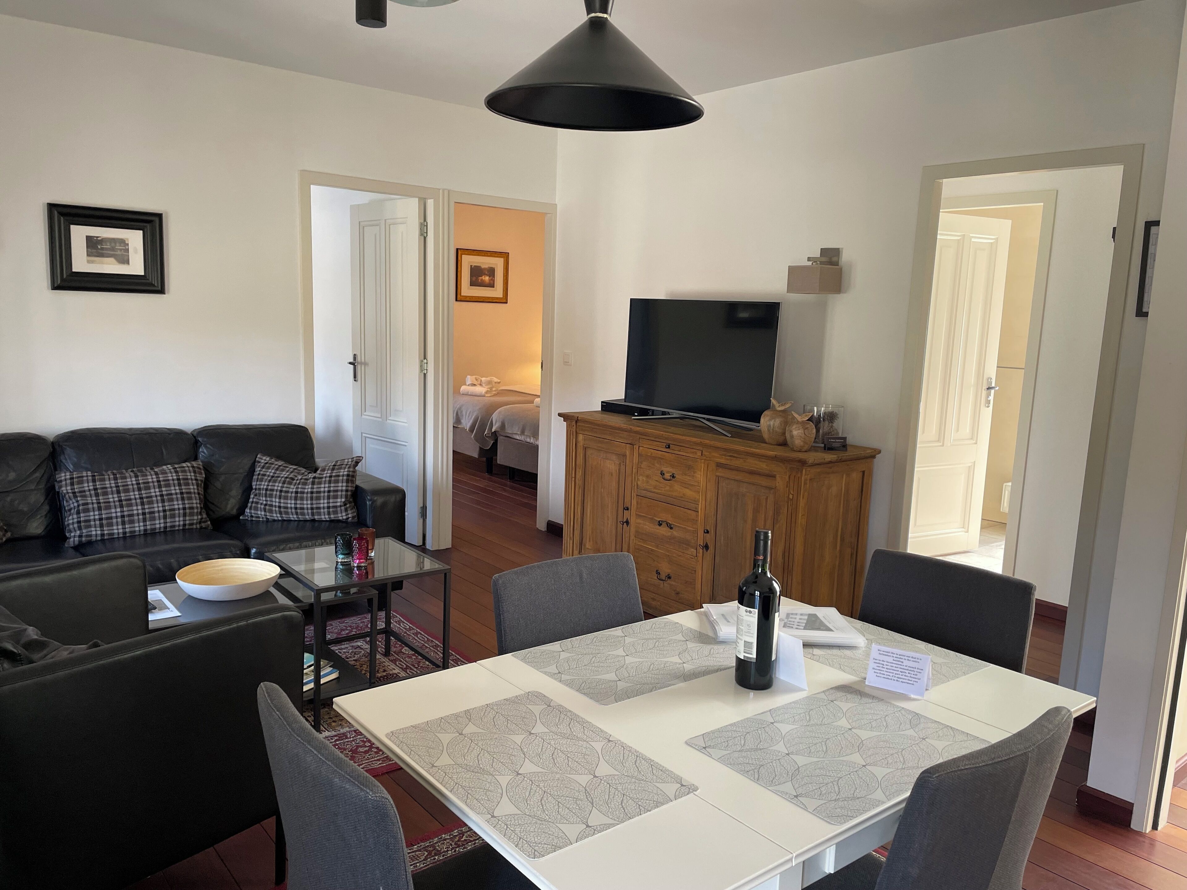 Ein Wohnzimmer mit Sofa, Couchtisch, Esstisch mit Stühlen, Fernseher auf einem Holzschrank und einem Flur, der zu einem Schlafzimmer führt.