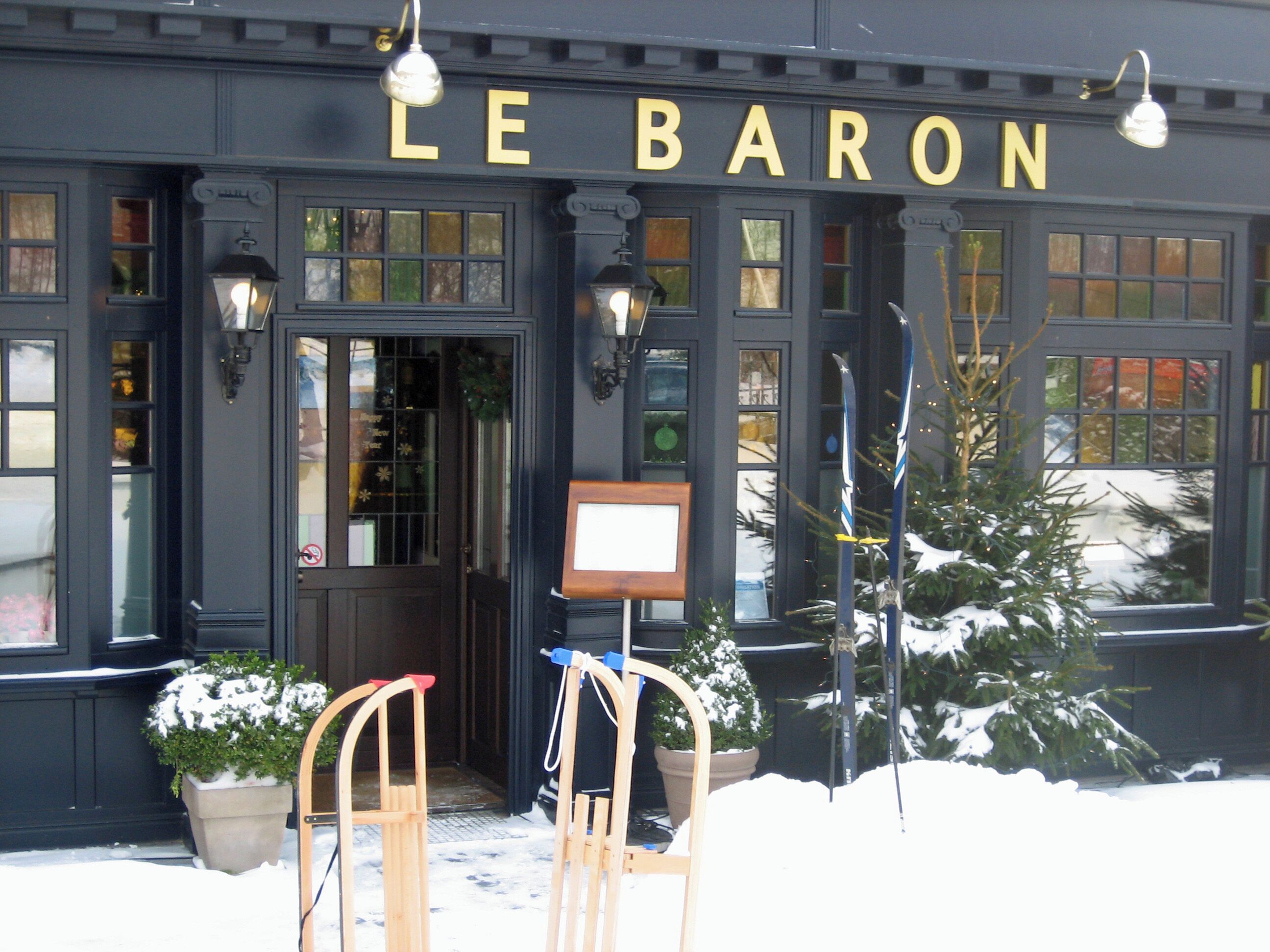 The exterior of Le Baron with a snow-covered ground, a wooden chair in front, and a Christmas tree to the right.