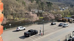 A parking lot with several cars, a river, and a hilly landscape with trees and buildings in the background. - Le Baron Suites & Apartments (Stavelot)