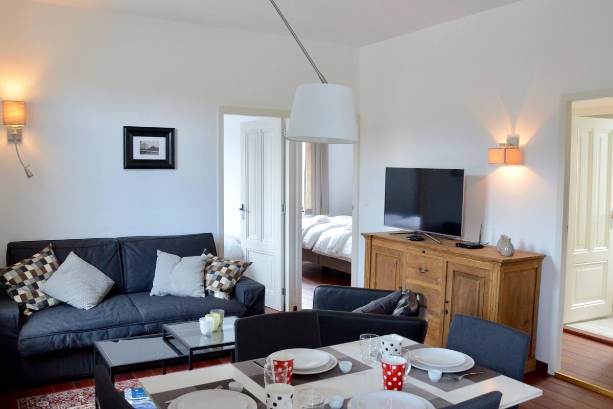 A modern living room with a sofa, coffee table, and dining area. A TV is mounted on the wall, and there is a wooden cabinet with a TV on top.