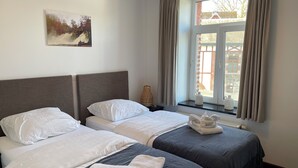 A hotel room with two beds, a painting on the wall, a window with curtains, and a view of buildings outside. - Le Baron Suites & Apartments (Stavelot)