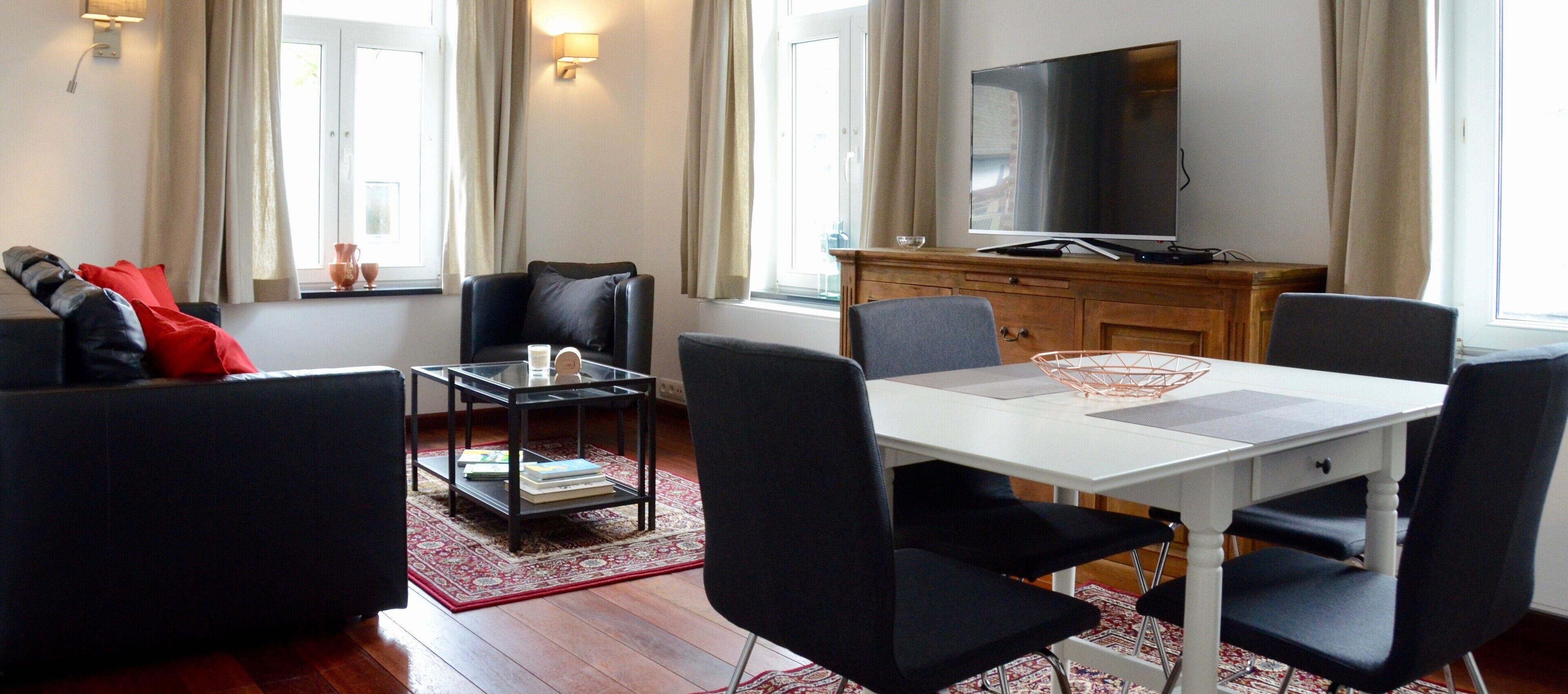 A modern living room with a white dining table, black chairs, a black sofa with red cushions, a glass coffee table, a wooden cabinet, and a flat-screen TV.