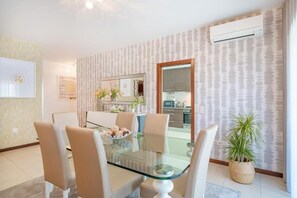 A modern dining area with a glass table and beige chairs, a potted plant, and a kitchen visible through an open door.