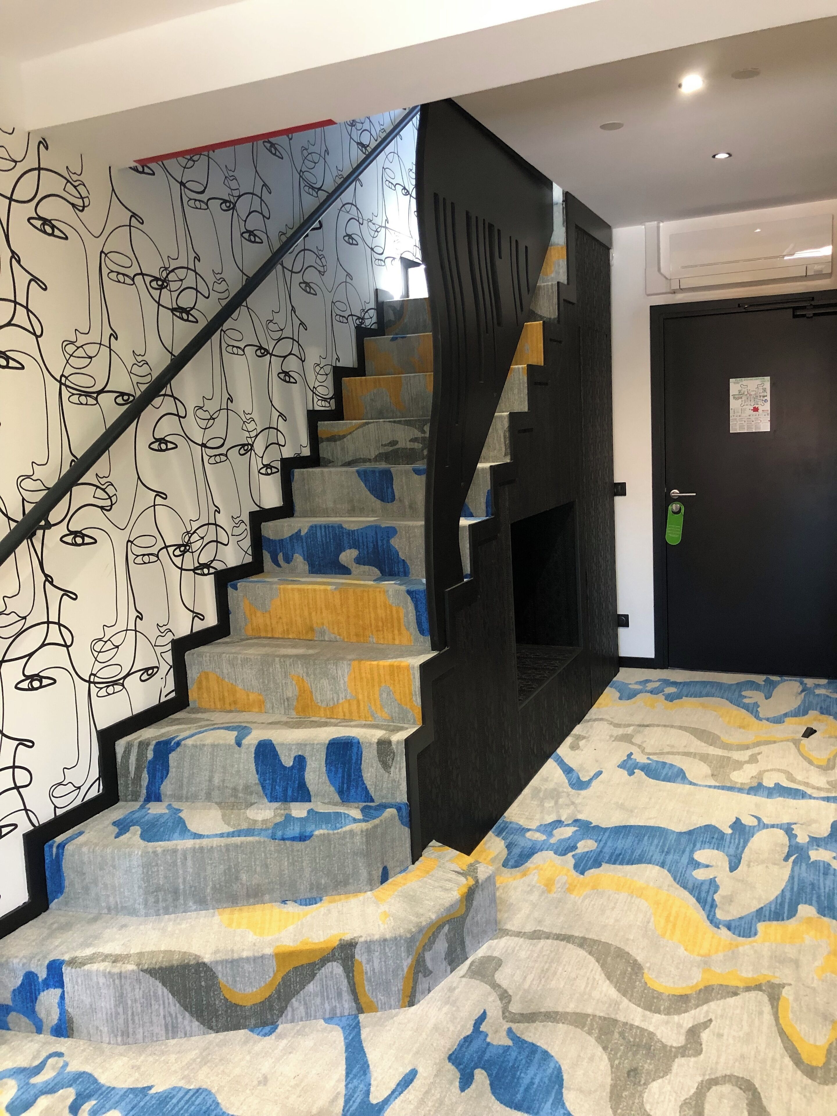 Una escalera con arte abstracto en la pared, una alfombra estampada y una puerta negra con una etiqueta verde.