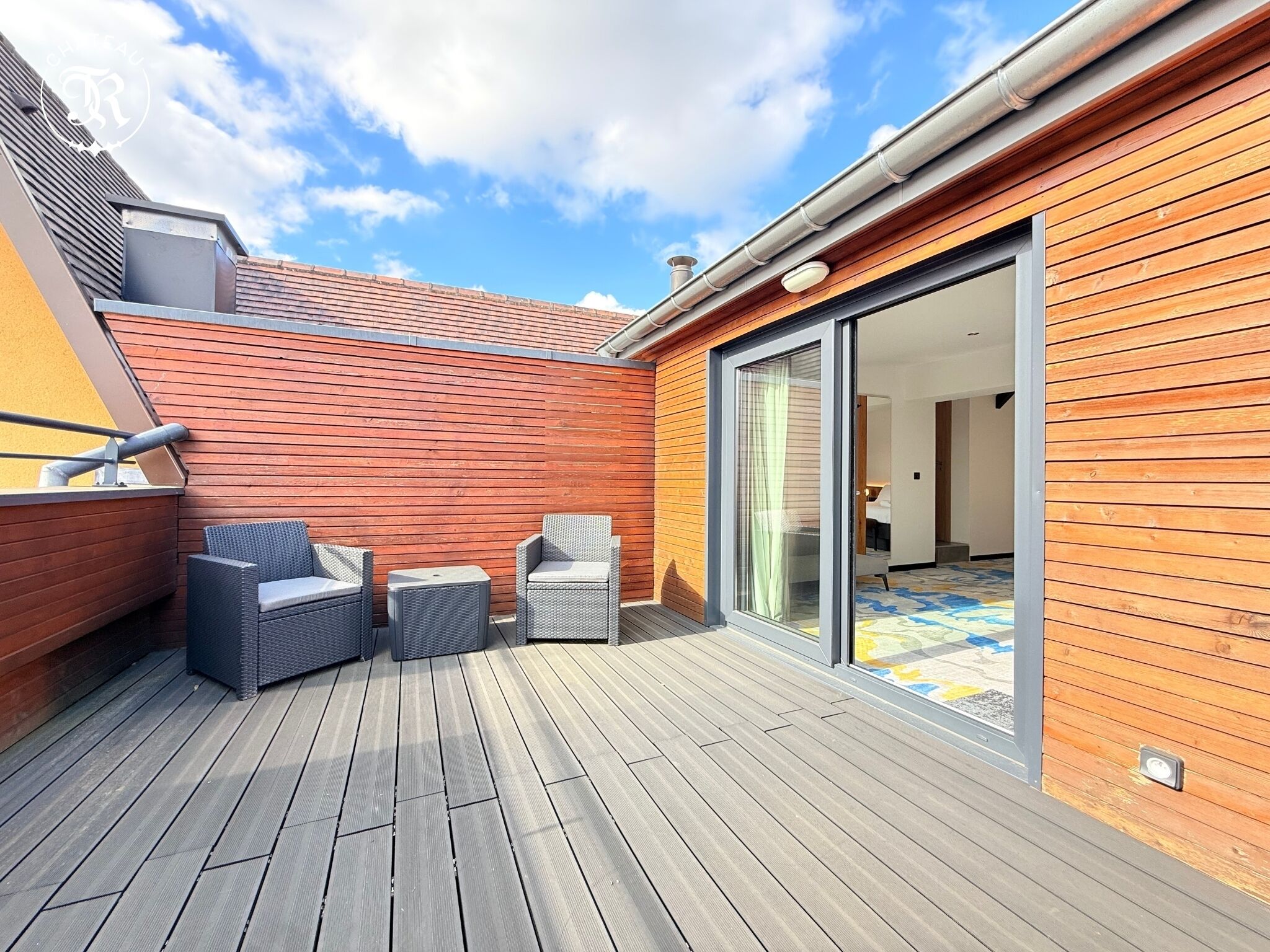 A wooden deck with two wicker chairs and a small table, a sliding glass door leading to an interior room, and a red wall with a metal roof.
