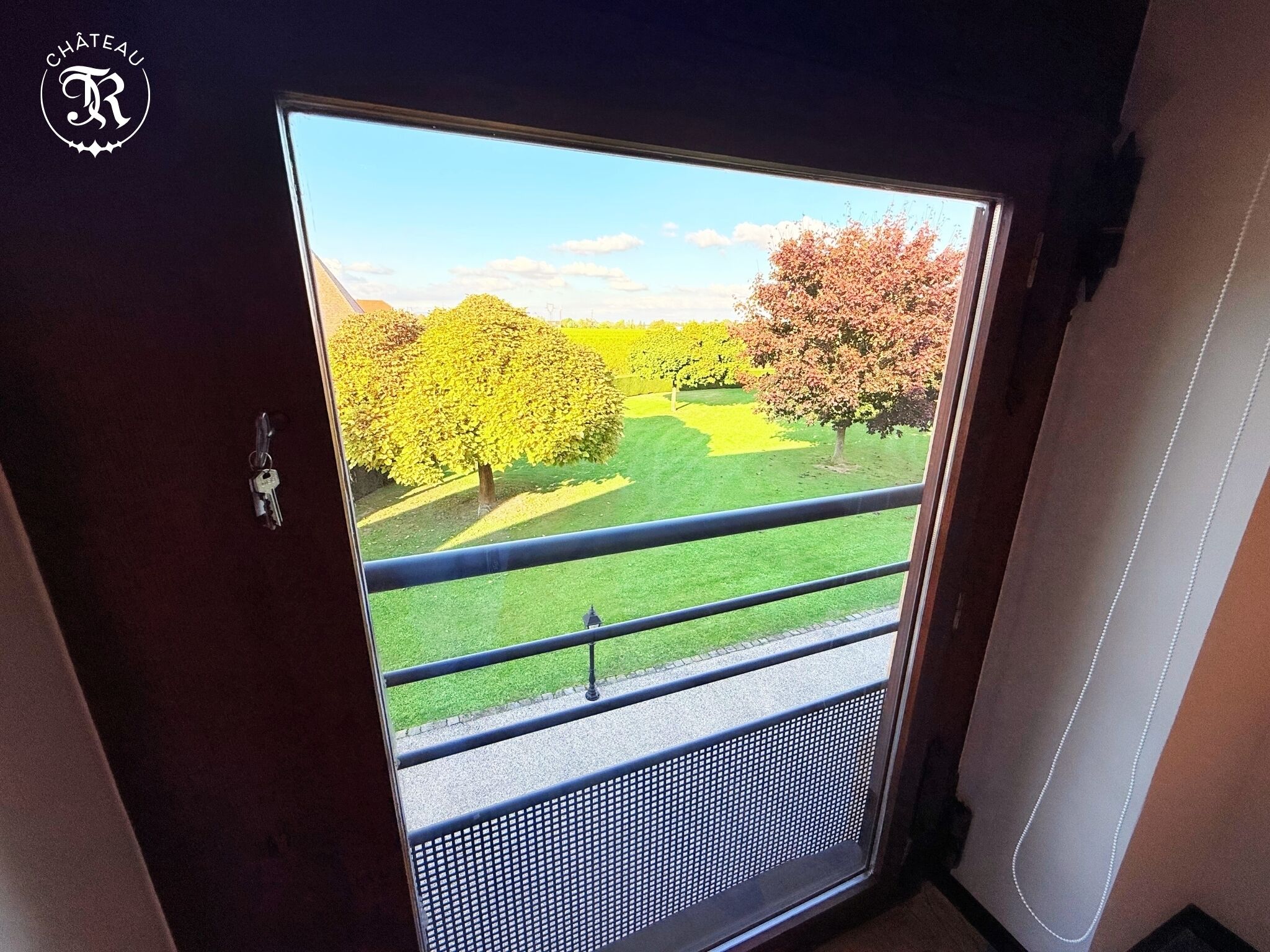 Vue depuis l’intérieur d’une pièce dotée d’une grande fenêtre donnant sur un jardin bien entretenu, avec des arbres et une pelouse.