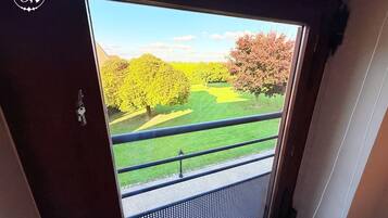 Vue depuis l’intérieur d’une pièce dotée d’une grande fenêtre donnant sur un jardin bien entretenu, avec des arbres et une pelouse.