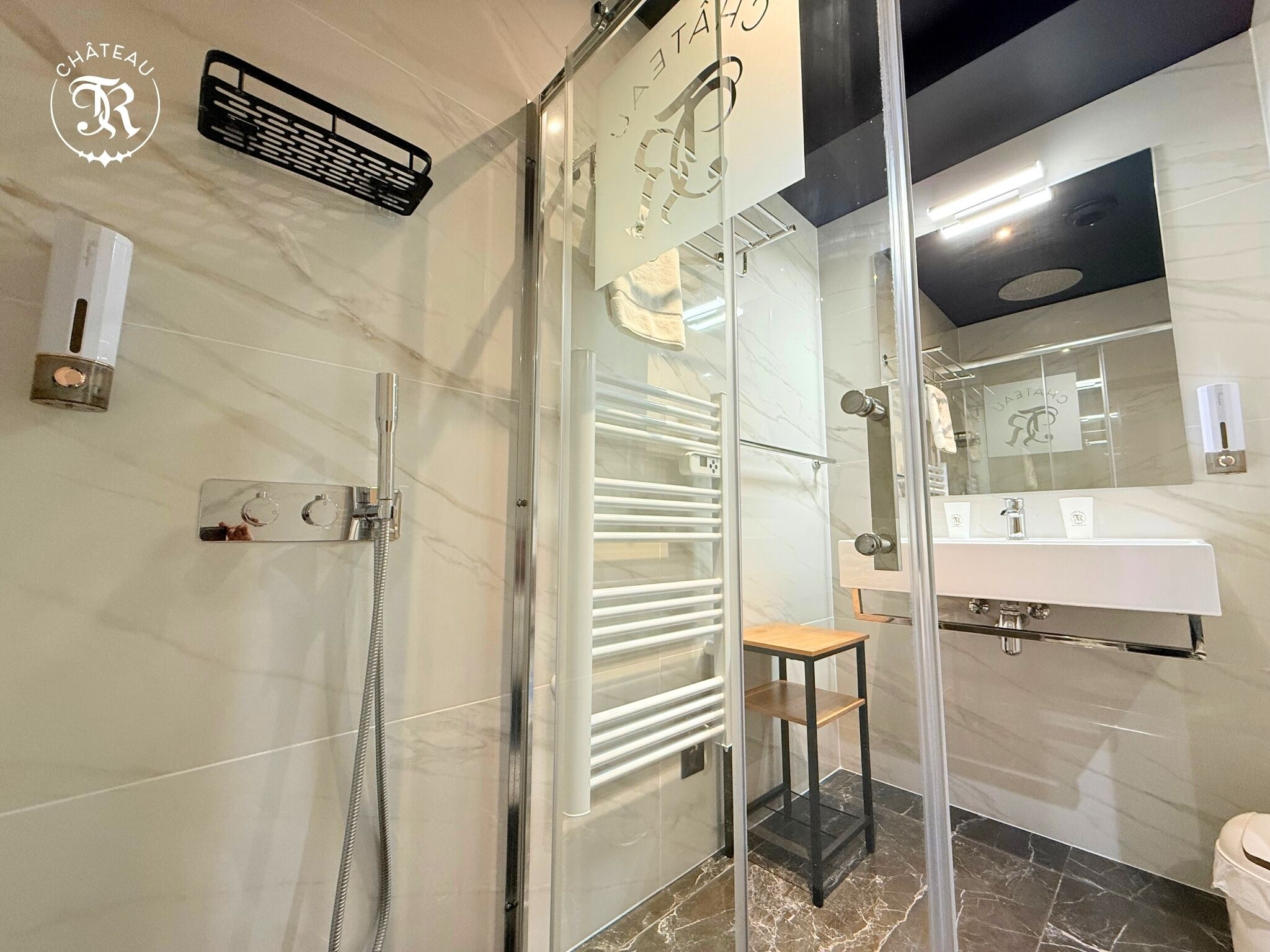 Une salle de bain moderne avec une douche à parois de verre, un lavabo blanc avec un tabouret en bois et un sol carrelé en marbre.