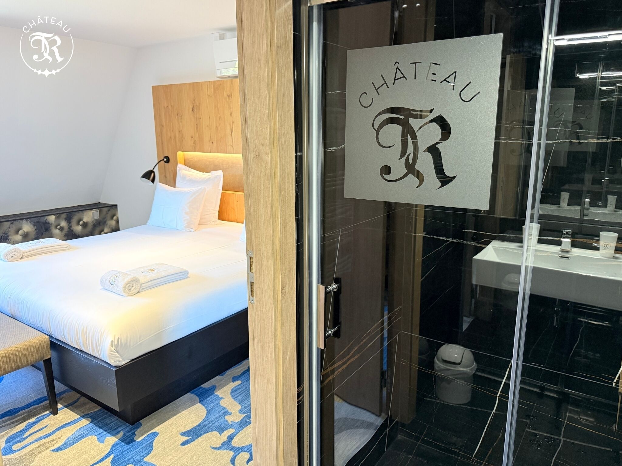 A hotel room with a bed, a bedside table, a lamp, a bathroom with a sink and mirror, and a sign reading "CHÂTEAU".
