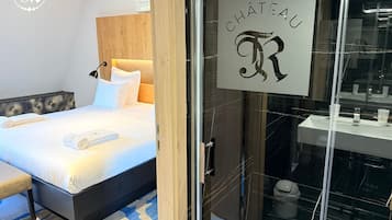 A hotel room with a bed, a bedside table, a lamp, a bathroom with a sink and mirror, and a sign reading "CHÂTEAU".