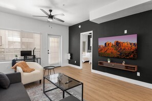 A modern living room with a ceiling fan, a flat-screen TV mounted on the wall, a sofa, a coffee table, and a painting of a desert landscape.