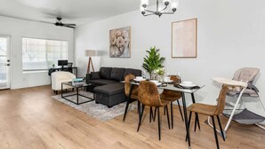 A modern living room with a sectional sofa, dining table, and chairs. There is a ceiling fan, a chandelier, and artwork on the walls.