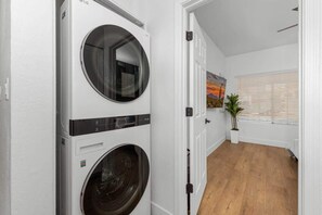 Un lavadero con dos lavadoras y secadoras apiladas, piso de madera, una puerta que da a una habitación con un cuadro, y una ventana con persianas.
