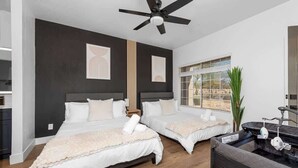 A modern bedroom with two beds, a ceiling fan, a window with blinds, and a small plant.