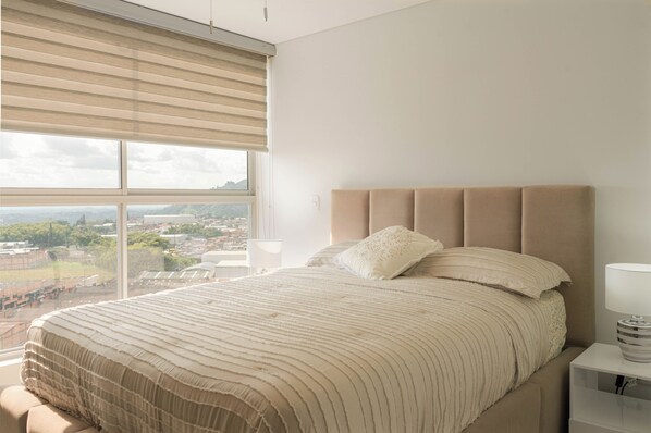 Une chambre avec une grande fenêtre, un lit doté d’une tête de lit beige et une lampe de chevet.