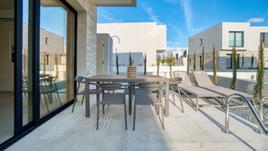 An outdoor patio with a table and chairs, a lounge area with sun loungers, and a view of modern buildings. - Avalon Gardens Villa 13 (Emba)