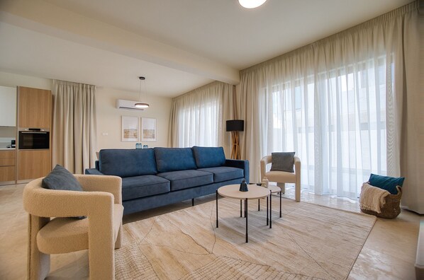 A modern living room with a sofa, armchairs, a coffee table, and a lamp. There is a kitchen area with an oven and a refrigerator. - Avalon Gardens Villa 13 (Emba)
