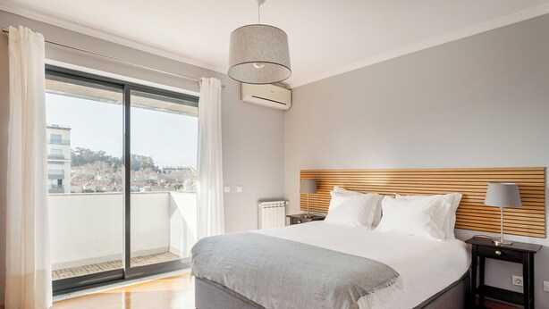 A modern bedroom with a large bed, a wooden headboard, a nightstand with a lamp, and a view of a cityscape through a sliding glass door. - Elegant Flat in Benfica (Lisbon)