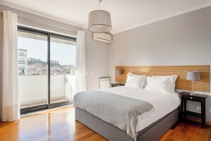 Un dormitorio moderno con una cama grande, un cabecero de madera, una mesita de noche con una lámpara y vistas a un paisaje urbano a través de una puerta corrediza de vidrio.