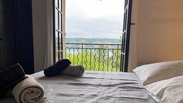 A bed with white linens and rolled towels, a view of greenery and hills through a balcony with a metal railing.