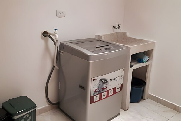 A compact, modern laundry machine with a control panel, a water inlet, and a small sink with a trash bin nearby. - Apto Sexta 103 (Pereira)