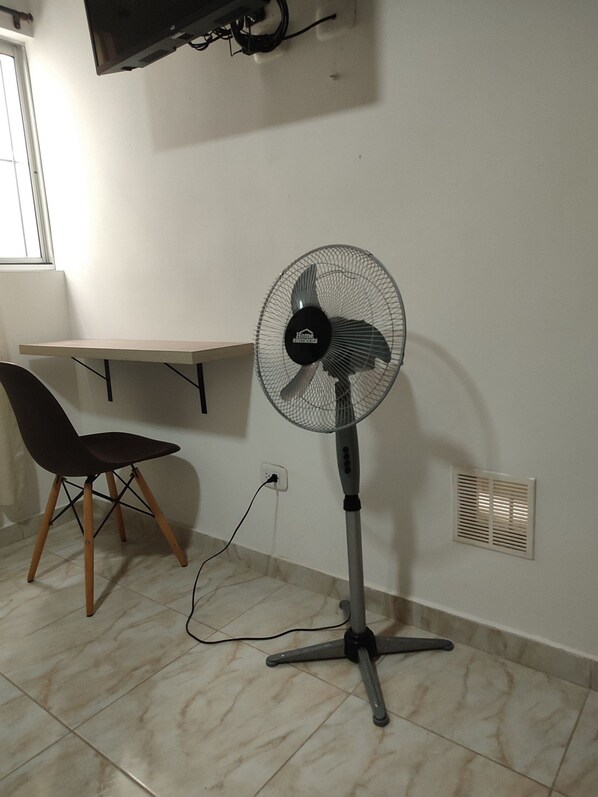 Une pièce équipée d’un ventilateur sur pied, d’une chaise, d’une table et d’un téléviseur fixé au mur.