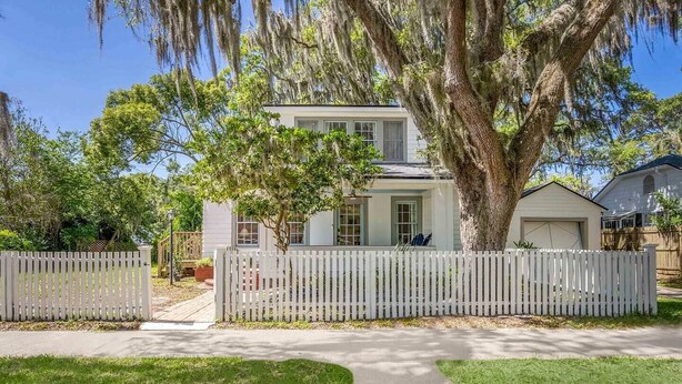 A two-story house with a white picket fence, a large tree in front, and a clear sky. - White Lily Cottage, White Lily Cottage (St. Augustine)