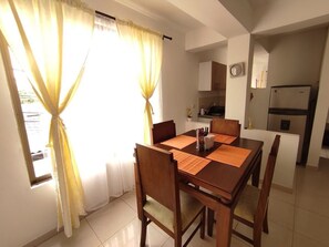 Zona pranzo con tavolo e sedie in legno, tende gialle e vista su una cucina con frigorifero.