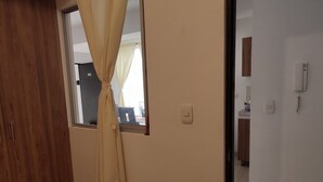 A room with a wooden door, a window with a curtain, a light switch, and a visible kitchen area in the background. - Jardin 201 (Pereira)