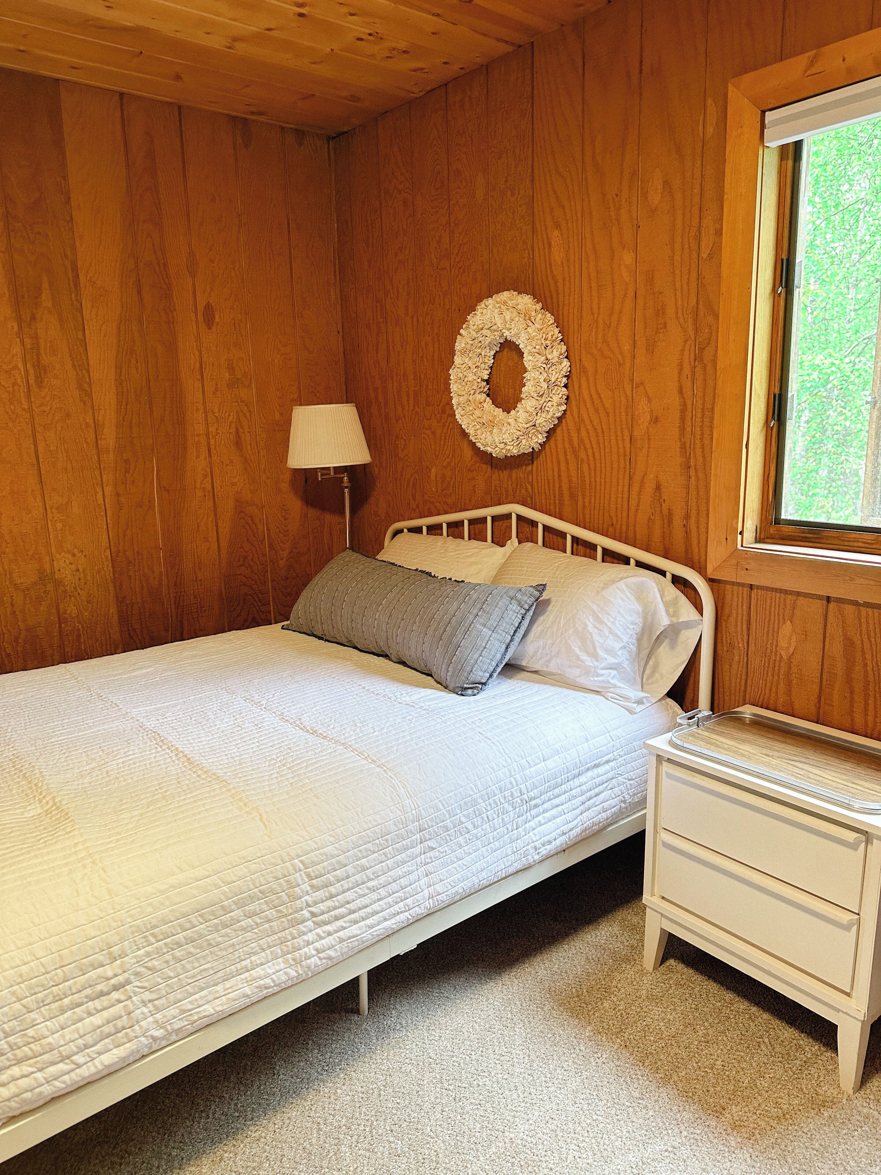 A wooden-paneled room with a bed, a nightstand, a lamp, and a window with a view of greenery.