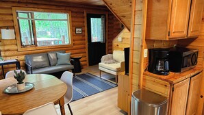 Interior acogedor de una cabaña con comedor, dos sofás, una cocina con cafetera y un cubo de basura.