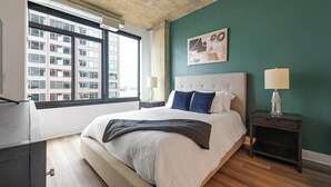 A modern hotel room with a large bed, a nightstand, a desk, and a window with a view of a building. - Landing | Spectacular 2BD, Gym, Game Room (Falls Church)