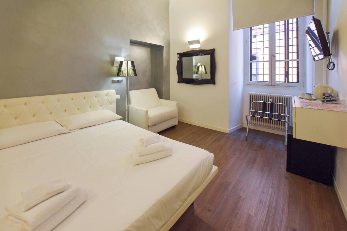 A hotel room with a large bed, a chair, a desk, a mirror, and a window with a view.
