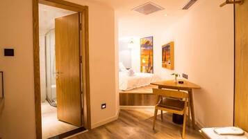 A hotel room with a wooden floor, a bed with white linens, a desk with a chair, and a small table with a lamp.