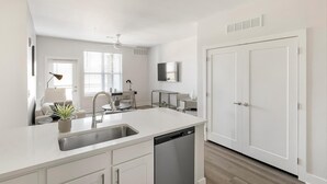 Una cocina moderna con encimera blanca, pileta de acero inoxidable y lavavajillas. Hay un comedor con una mesa y sillas, y un ventilador de techo.