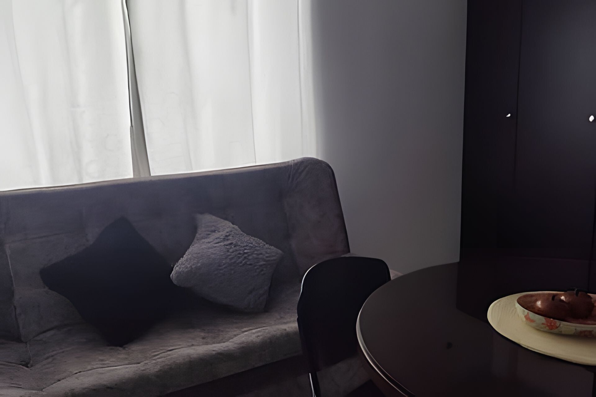 Una habitación con un sofá gris, una silla negra y una pequeña mesa redonda con un bol y un plato.