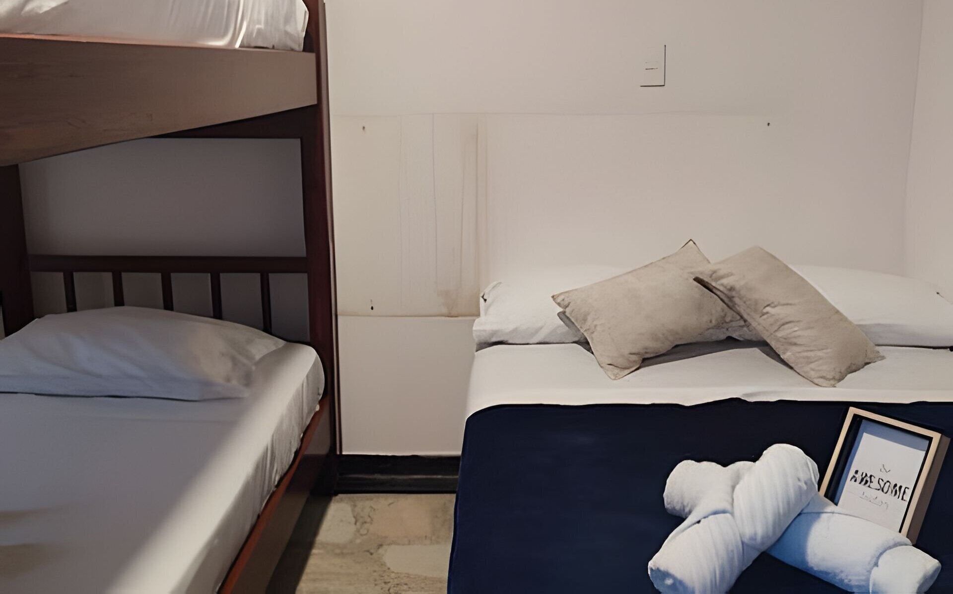 A room with two beds, one with a wooden headboard and the other with a plain headboard. There are two pillows on the lower bed, and a framed picture with the text "A BE SOME" on the floor.