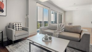 Ein modernes Wohnzimmer mit einem grauen Sofa, einem kleineren Sessel und einem Couchtisch. Ein großes Fenster bietet Blick ins Freie.