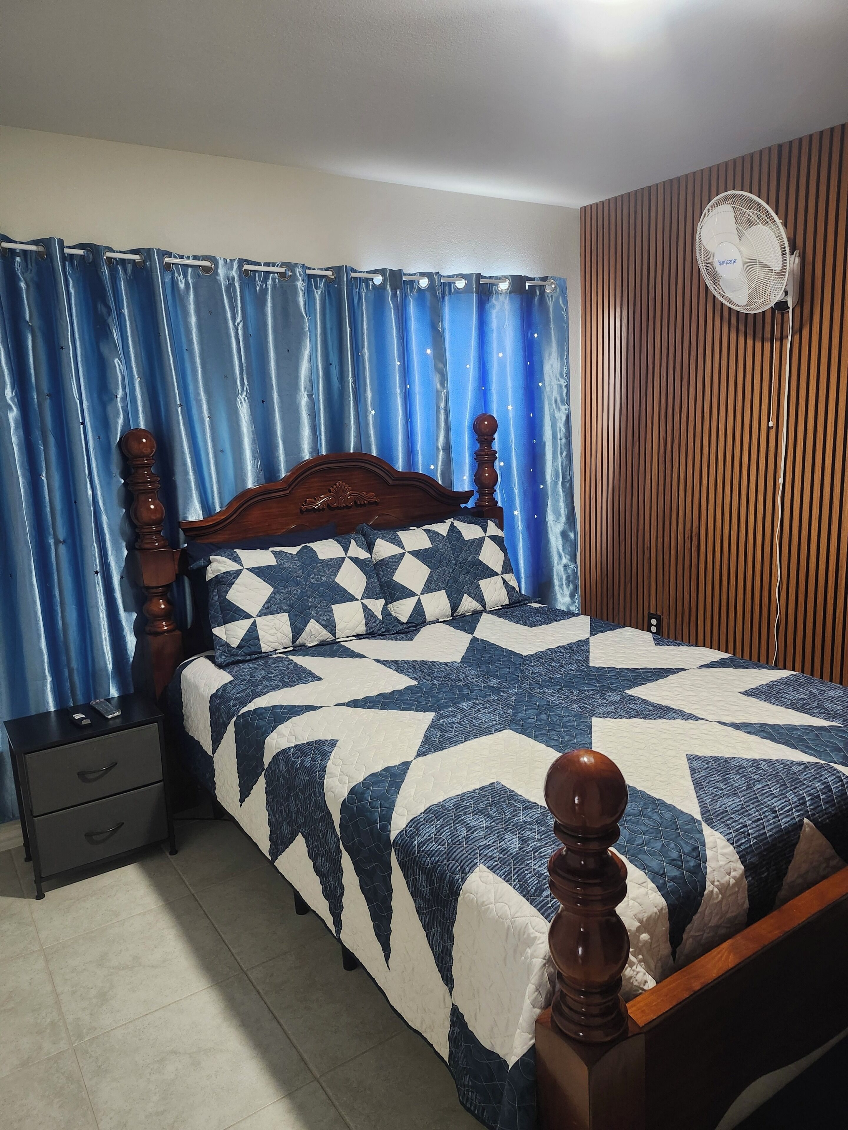 A bedroom with a wooden bed frame, a blue and white quilt, a nightstand with drawers, a wall-mounted fan, and a curtain with a star pattern.