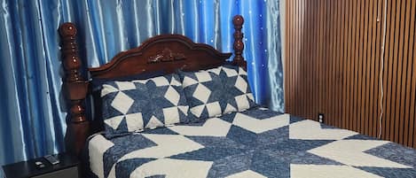 Une chambre à coucher avec un cadre de lit en bois, une couette bleue et blanche, une table de chevet avec des tiroirs, un ventilateur fixé au mur et un rideau orné de motifs en forme d’étoiles.