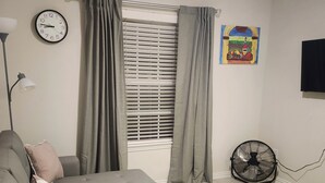 Ein Wohnzimmer mit einem Ecksofa, einer Wanduhr, einem Fenster mit Jalousien, einem Ventilator und einem Gemälde.