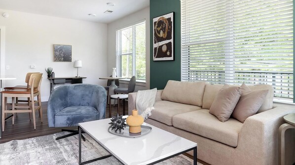 A modern living room with a beige sofa, a blue armchair, and a white coffee table. There is a dining area with a table and chairs, and artwork on the walls. - Landing | Amazing 2BD, Pool, Game Room (Atlanta)