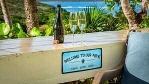 Una mesa con una botella y dos copas de vino blanco, un letrero que dice 'Bienvenidos a nuestro patio', y una vista panorámica del océano y las colinas al fondo.